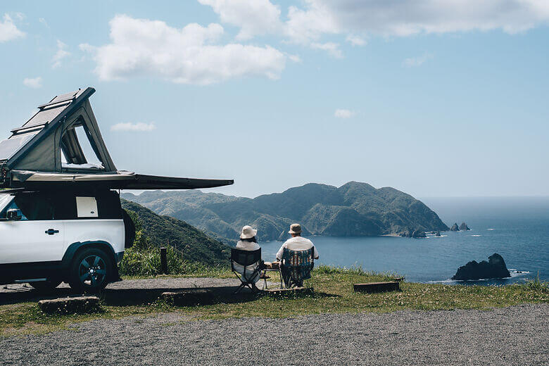 Zwei Menschen sitzen neben dem Camper und schauen auf eine Bucht im Süden von Japan